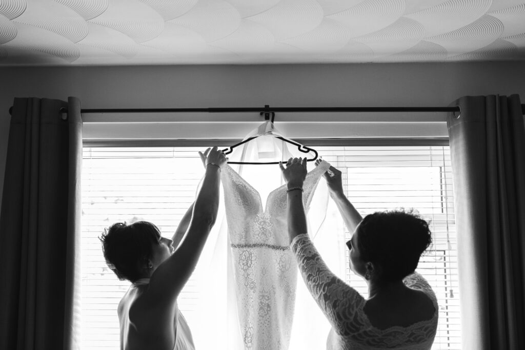 Mother of the bride and masid of honour reach for wedding dress hung in the window 