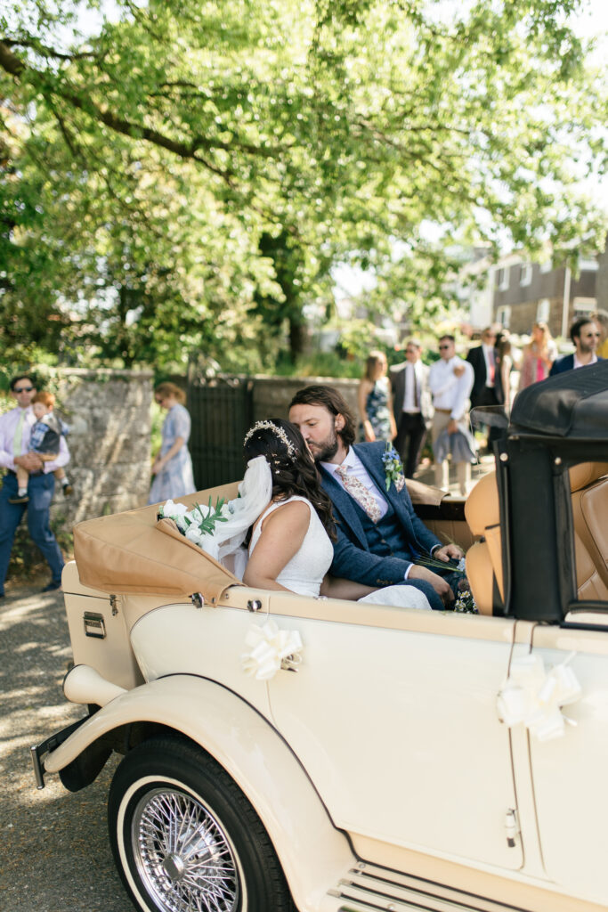 Bride and groom kiss in the back of the open top car, outside of the church they got married 