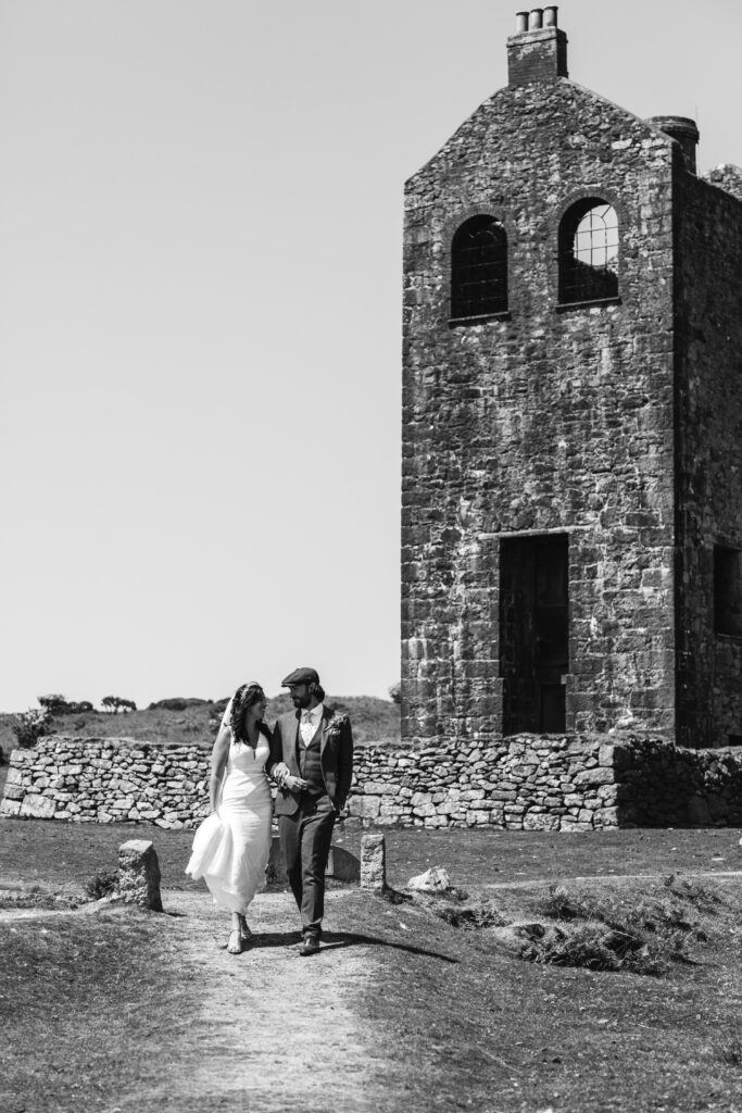 Couple walk towards the camera holding hands with an old Cornish mine in the background