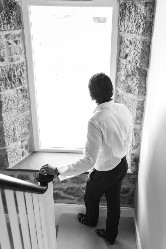 Groom looks out of the window as he walks down the stairs to get ready for his wedding day