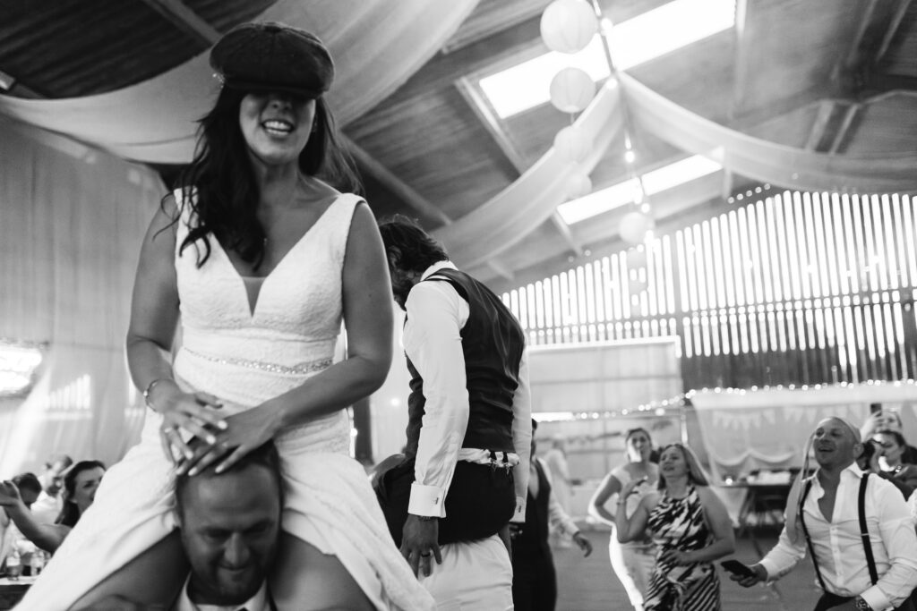 Bride sits on a mans shoulders and wears his hat on wedding day during the dancefloor 